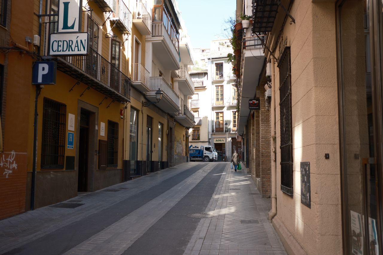 Home Explore a quiet narrow street in Granada, flanked by picturesque buildings and a calm atmosphere.